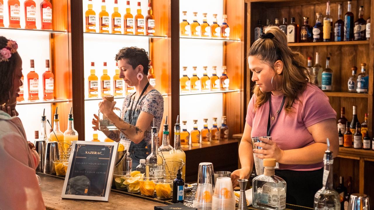 Two bartenders pouring cocktail creations in front of a display bottle wall at the Sazerac House